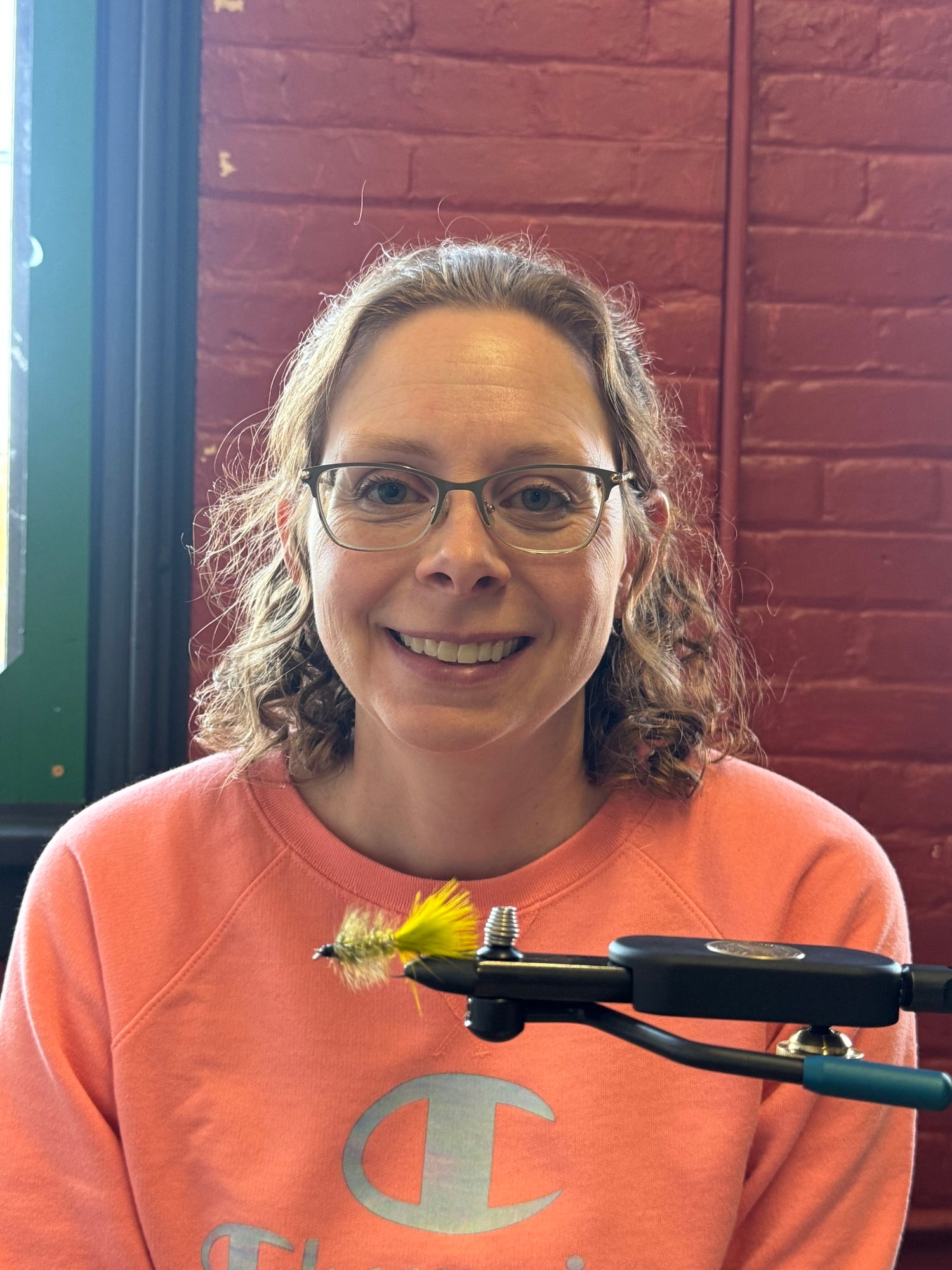 Person wearing glasses and an orange sweatshirt with a fly fishing tool in front of a brick wall.