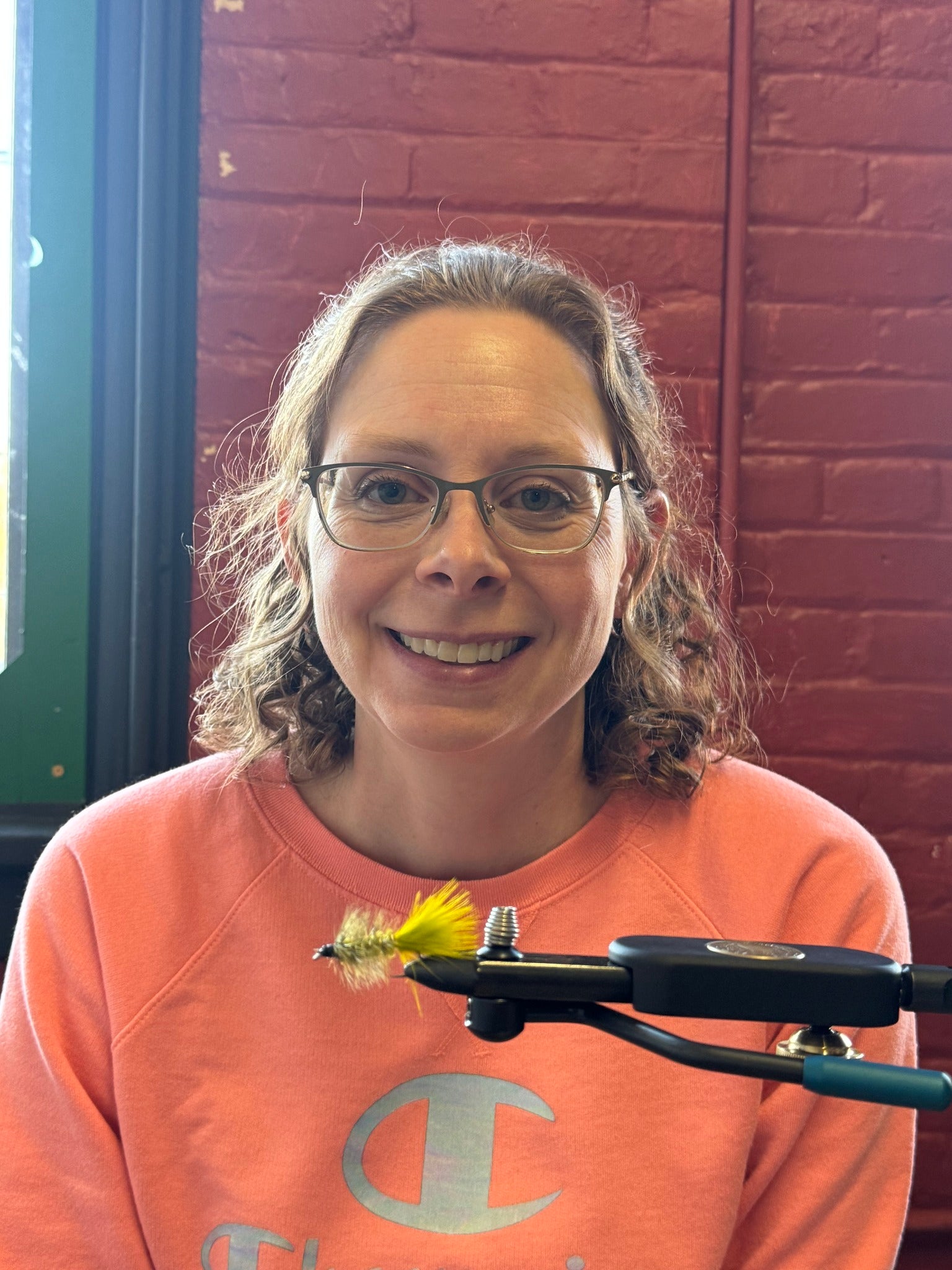 Person wearing glasses and an orange sweatshirt with a fly fishing tool in front of a brick wall.