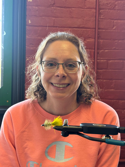 Person wearing glasses and an orange sweatshirt with a fly fishing tool in front of a brick wall.